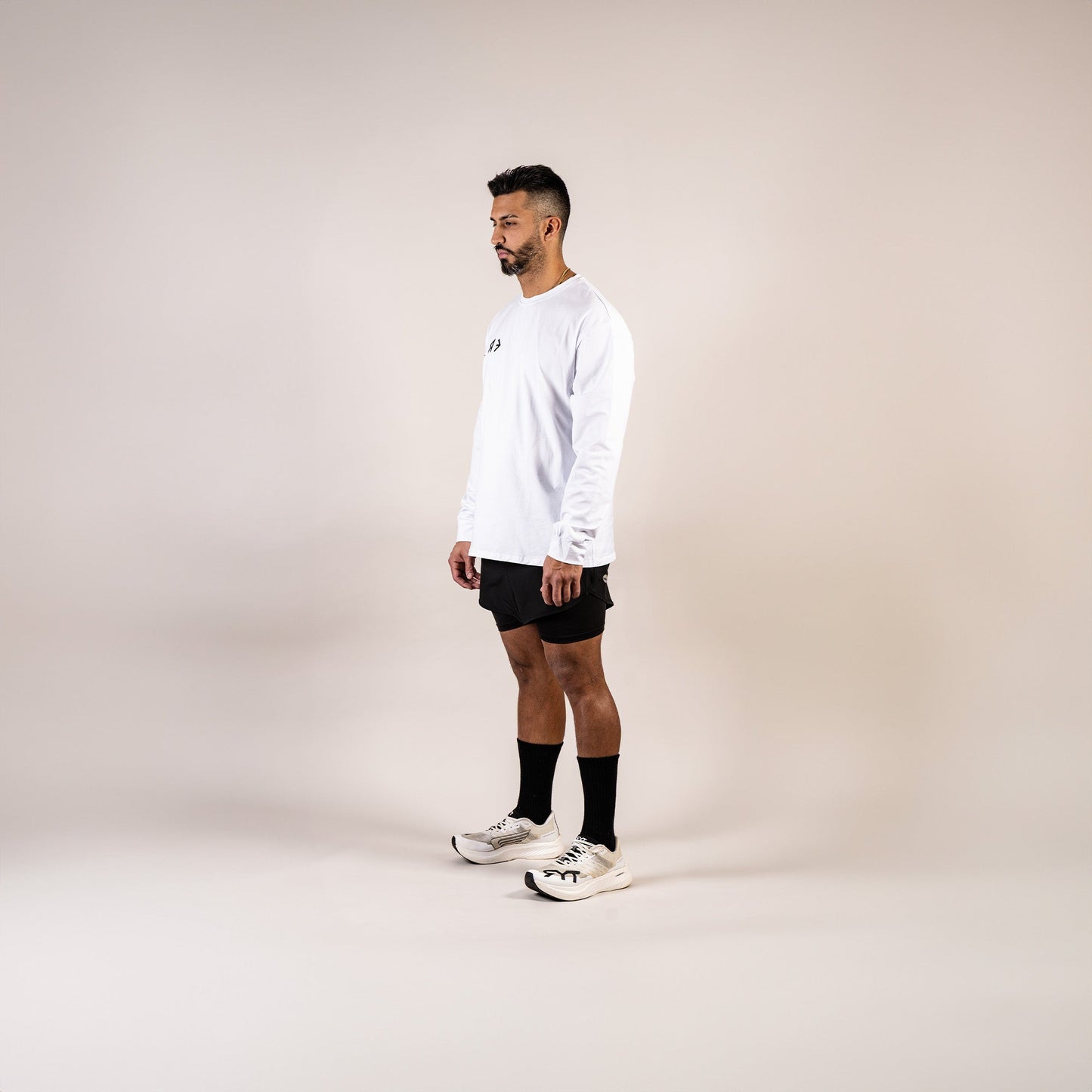 Man wearing a white long-sleeve shirt, black shorts, and white sneakers on a beige background