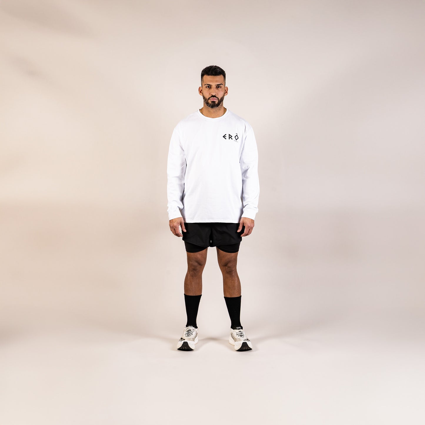 Man wearing a white long-sleeve shirt, black shorts, and white sneakers on a beige background