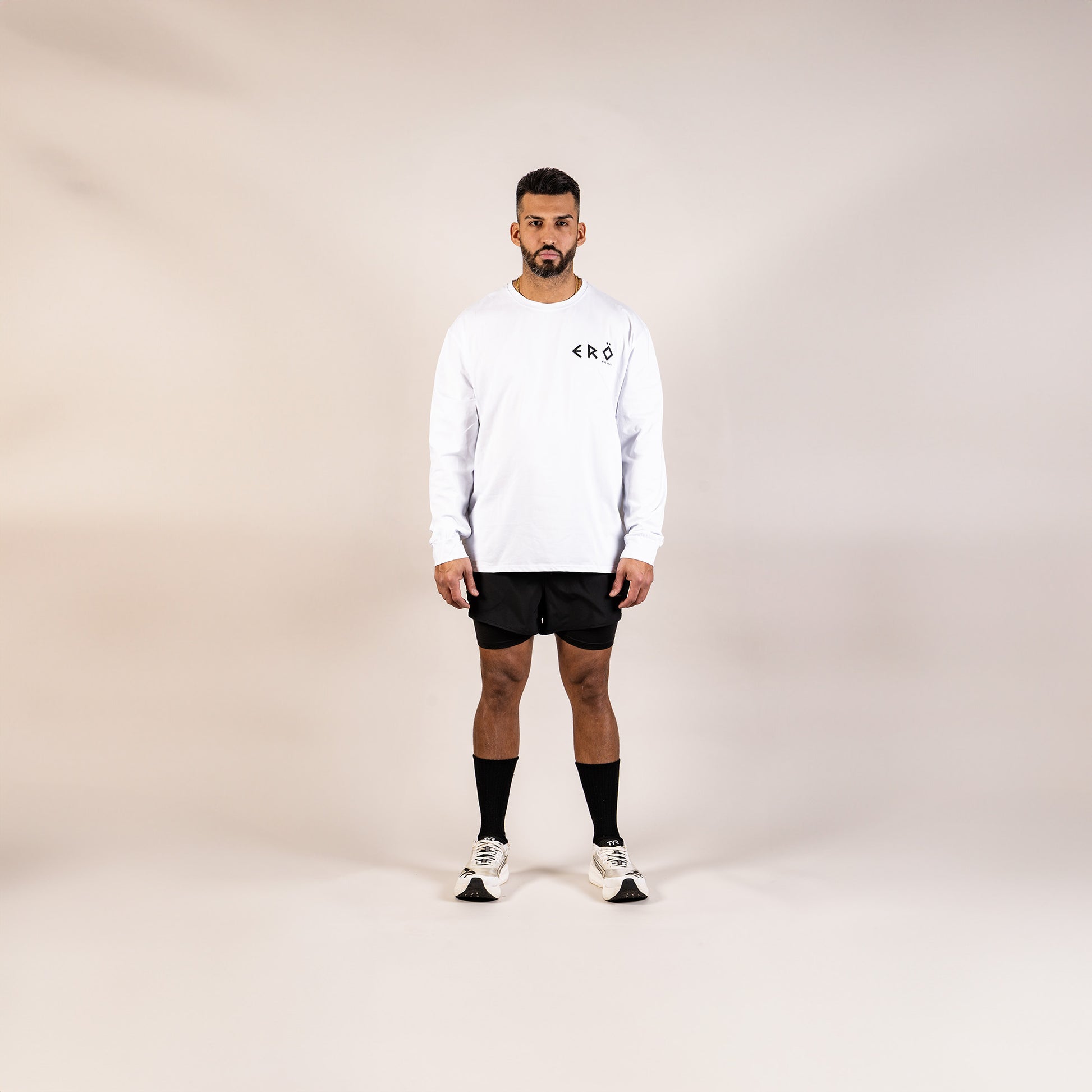 Man wearing a white long-sleeve shirt, black shorts, and white sneakers on a beige background