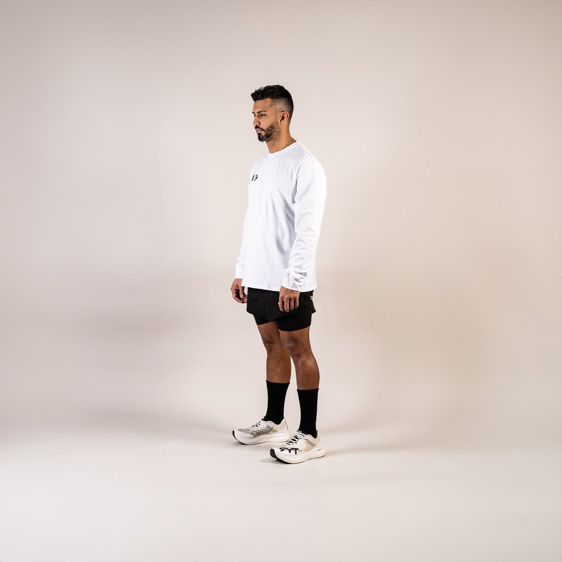 Man wearing a white long-sleeve shirt, black shorts, and white sneakers on a beige background