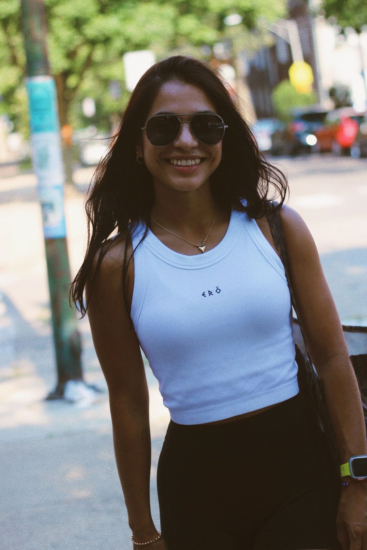 Woman wearing a light blue tank top with a brand logo, sunglasses, and carrying a bag on a city street.