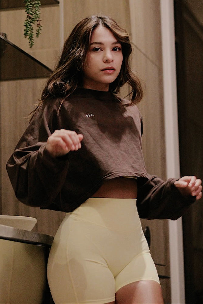 Woman wearing a brown cropped sweatshirt and light yellow shorts in a bathroom.