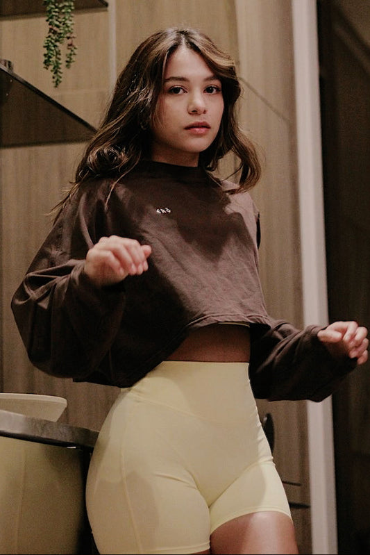 Woman wearing a brown cropped sweatshirt and light yellow shorts in a bathroom.