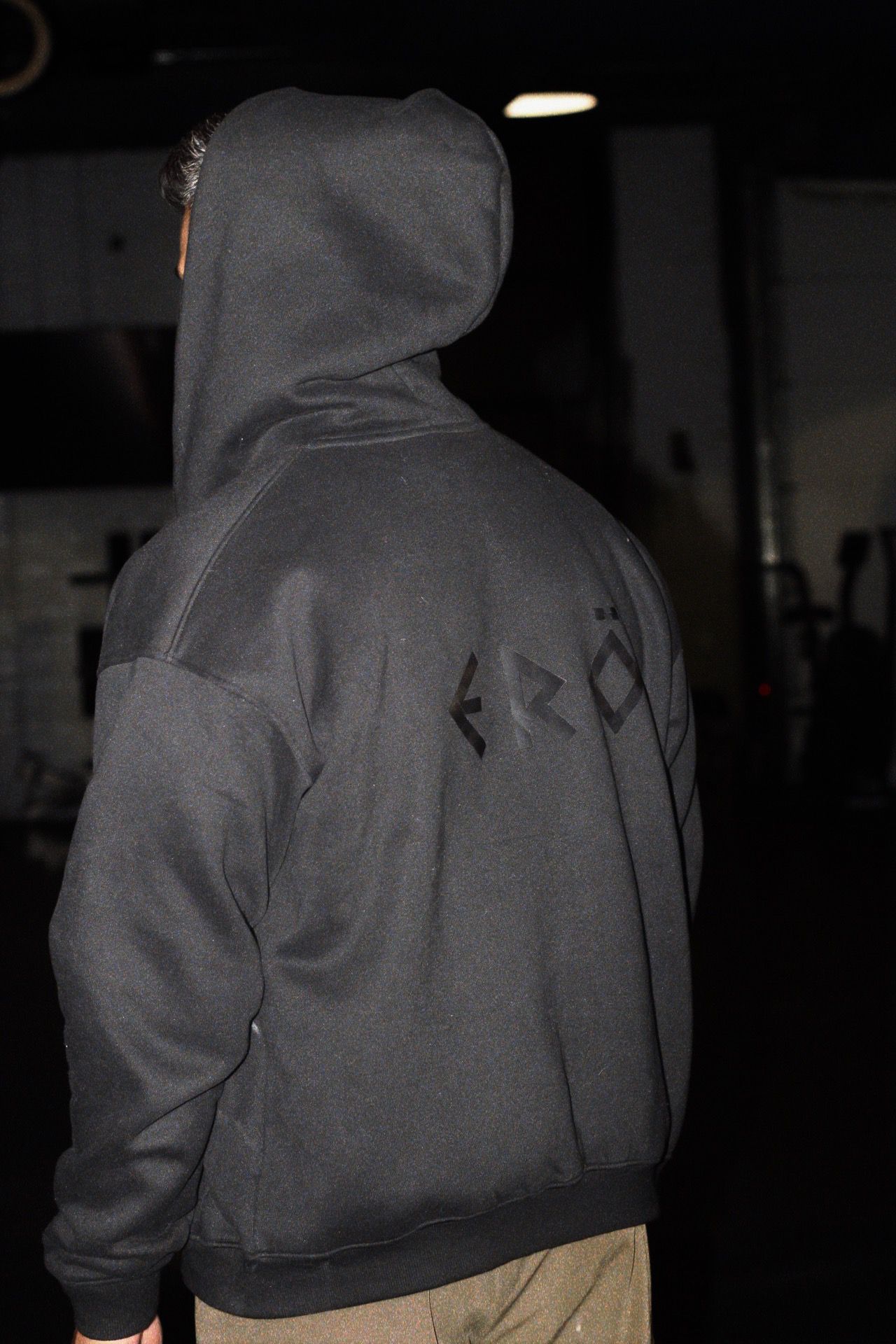 Person wearing a gray hoodie with a brand logo on the back, standing in a dark indoor setting.